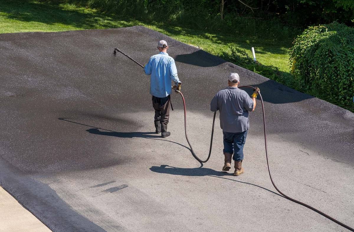 Two workers spray-applying black sealer to an asphalt driveway