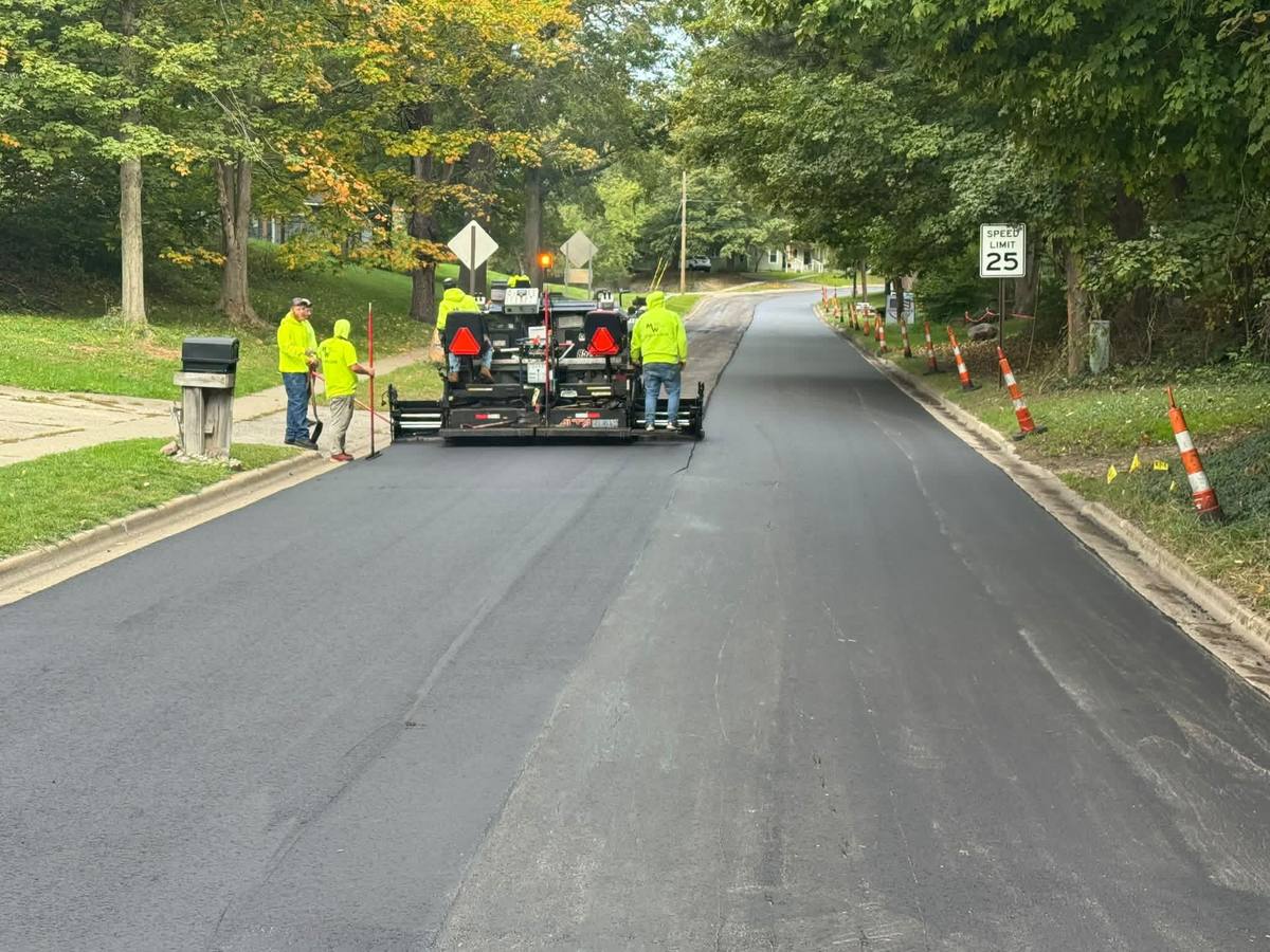 Paving crew in high-visibility shirts laying asphalt with a paver on a tree-lined road